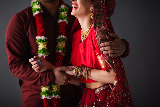 cropped view of indian man in floral garland hugging happy bride in traditional headscarf isolated on grey  - 写真・画像