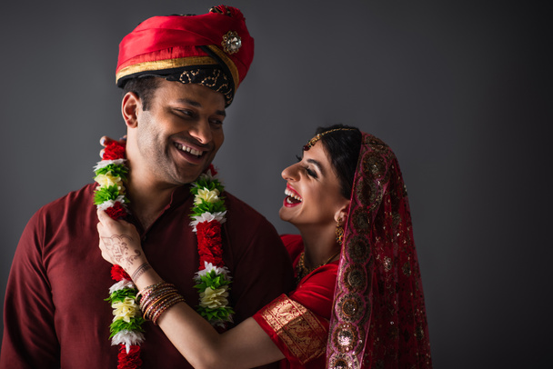 fröhliche indische Braut trägt Girlande auf Bräutigam isoliert auf grau  - Foto, Bild