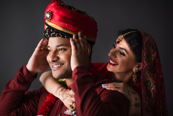 χαρούμενη Ινδή νύφη αγκαλιάζει το γαμπρό στο τουρμπάνι απομονωμένη στο γκρι  - Φωτογραφία, εικόνα