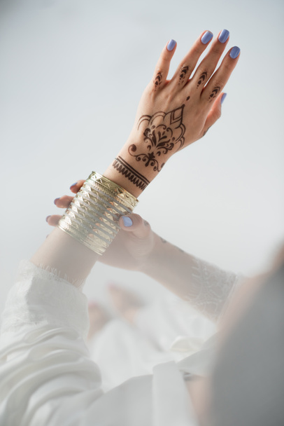 vista parcial de la mujer india con mehndi usando pulsera de oro mientras se prepara para la boda en blanco - Foto, imagen