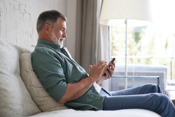 Smiling Senior Man Using Cellphone Browsing Internet Sitting On Sofa Indoor. - 写真・画像