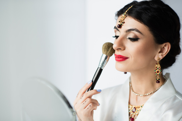 indian bride with mehndi applying face powder with cosmetic brush near mirror on white  - Photo, image