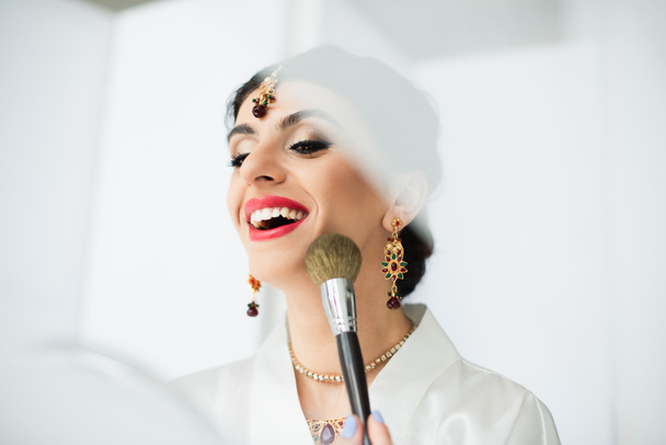 cheerful indian bride applying face powder with cosmetic brush on white  - Photo, image