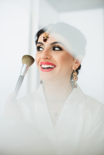 radosna indyjska panna młoda nakładająca proszek do twarzy pędzlem kosmetycznym na biały  - Zdjęcie, obraz