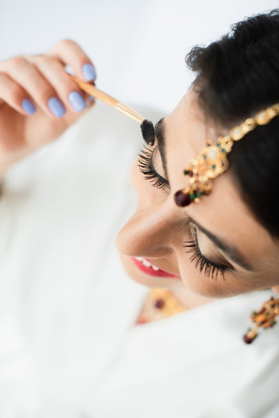 Draufsicht der glücklichen indischen Braut mit mehndi Styling Augenbraue mit Kosmetikpinsel auf weiß  - Foto, Bild