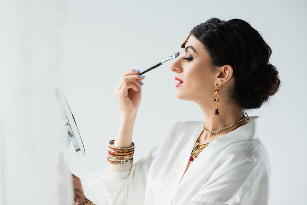 vue latérale de la mariée indienne avec mehndi appliquer ombre à paupières avec pinceau cosmétique sur blanc  - Photo, image