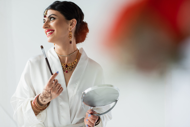 joyful indian bride with mehndi holding mirror and cosmetic brush on white  - Fotoğraf, Görsel
