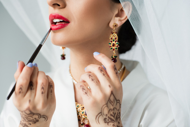 cropped view of indian bride applying lipstick with cosmetic brush on white - Zdjęcie, obraz