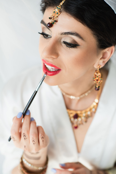 Novia india aplicando lápiz labial con pincel cosmético sobre blanco - Foto, imagen