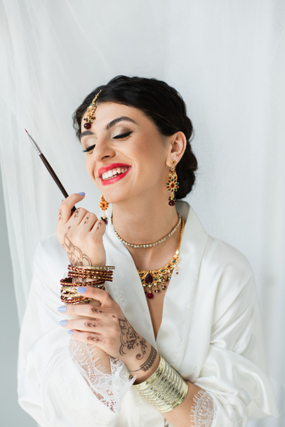 pleased indian bride with mehndi holding cosmetic brush on white  - Φωτογραφία, εικόνα