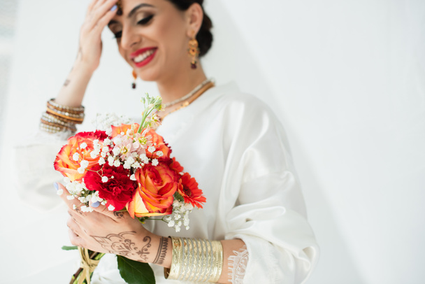 young blurred indian bride with mehndi smiling while holding bouquet of flowers on white - Fotografie, Obrázek