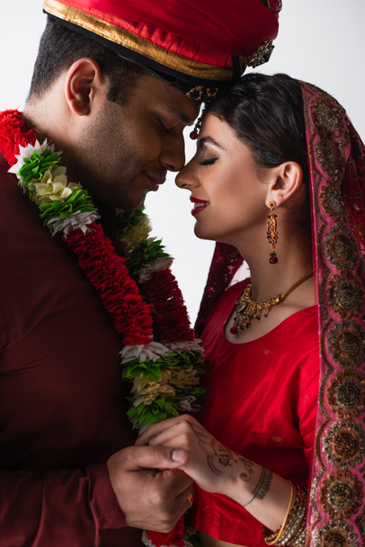 Seitenansicht eines lächelnden indischen Ehepaares in traditioneller Kleidung, das die Hände isoliert auf Weiß hält - Foto, Bild