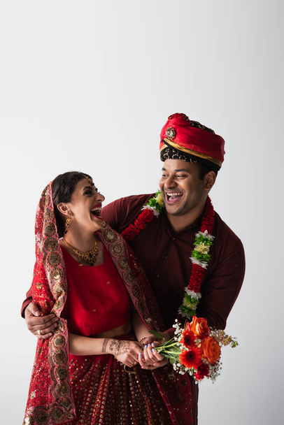 Ευτυχισμένος Ινδός γαμπρός στο τουρμπάνι αγκαλιάζει χαρούμενη νύφη στο σάρι με λουλούδια απομονωμένα στο γκρι - Φωτογραφία, εικόνα