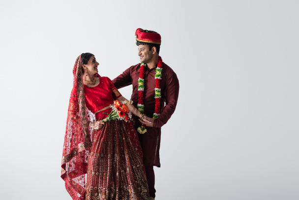 χαμογελαστός Ινδός άνδρας με τουρμπάνι κοιτάζοντας όμορφη νύφη στο σάρι με μπουκέτο λουλούδια απομονωμένα σε λευκό - Φωτογραφία, εικόνα