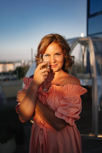 at sunset. beautiful woman in a pink dress on the roof with a glass of champagne - Фото, изображение