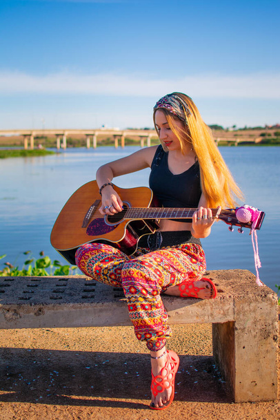 かなりブロンドのアルゼンチン人女性がベンチに座って川沿いでギターを弾いて - 写真・画像