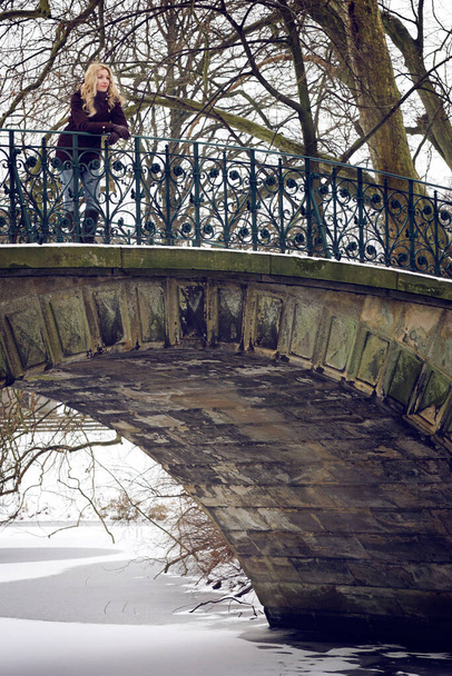 A vertical shot of a young Caucasian female standing on a bridge in a snowy park - Zdjęcie, obraz