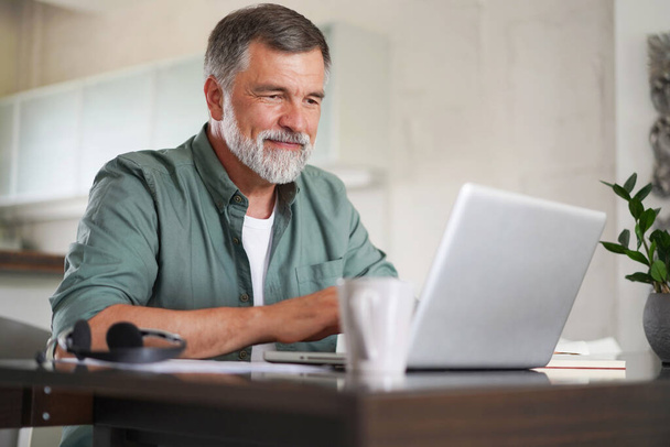 Handsome mature man in casual suit sitting at the table in home office and working at laptop - Foto, Imagen