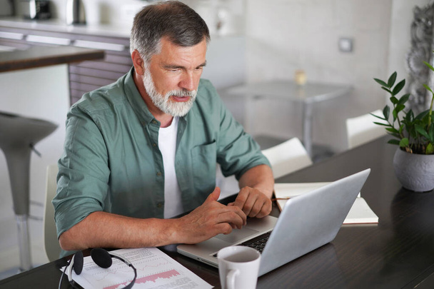 Handsome mature man in casual suit sitting at the table in home office and working at laptop - Photo, image