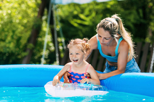 Молода мама вчить свою маленьку доньку плавати в басейні в саду за допомогою надувного кола
 - Фото, зображення