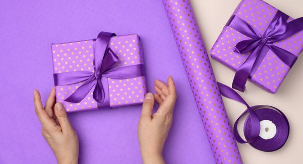 two female hands are holding purple gift box on paper background, concept of congratulations on birthday, top view - Photo, Image