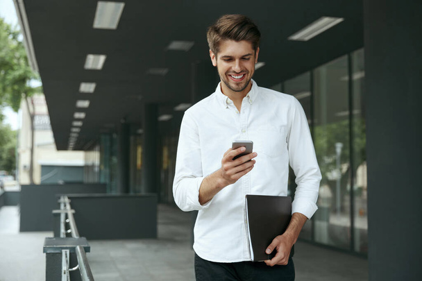 Pleased european businessman using mobile phone and holding documents. Young man standing on background of modern office building in city. Smiling bearded guy wear white shirt and jeans. Daytime - Fotó, kép