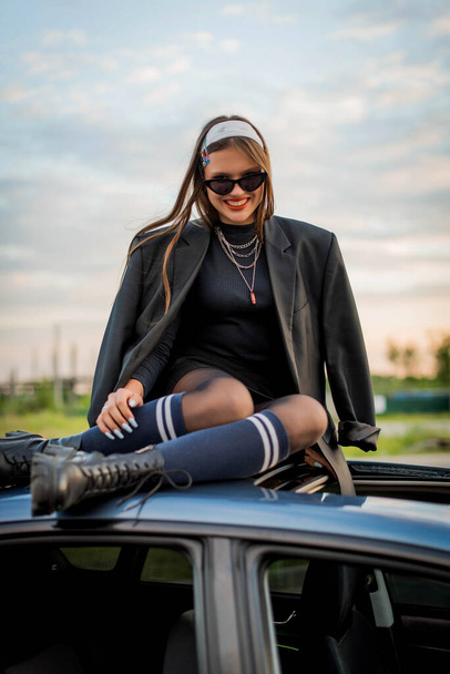 Beautiful country girl in 90s style on the roof of the car Back in time 90s 80s. Stylish girl in retro colourful vintage coat. dressed in street style clothes with chains around neck for a walk. Youth and lifestyle concept - Photo, Image