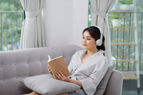 Una mujer está leyendo un libro y escuchando música mientras se sienta en un sofá gris. - Foto, imagen