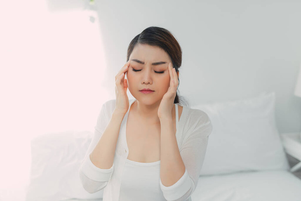 Mladá žena s bolestí hlavy, sedí na posteli a dotýká se spánků - Fotografie, Obrázek