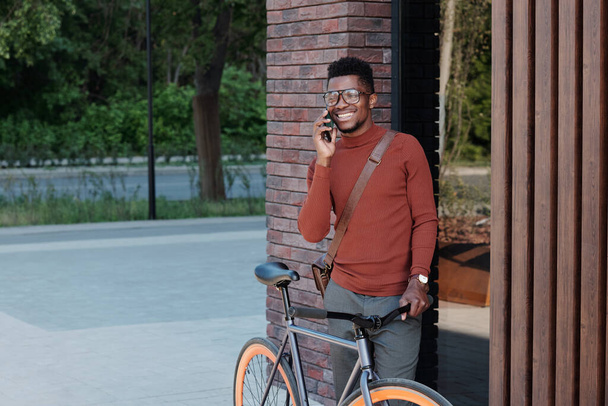 Junger afrikanischer Geschäftsmann telefoniert mit Fahrrad durch modernes Gebäude - Foto, Bild