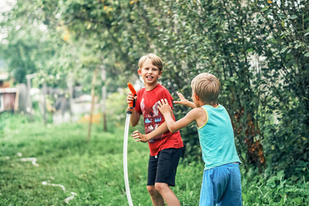 Joyful funny boy with cute little brother plays with water hose in green garden in spacious summerhouse yard on nice sunny day - Photo, image