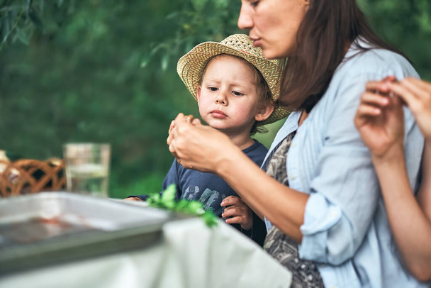 Young mother and little son in straw hat with food sit at table during picnic in green park on nice summer day close view - Fotoğraf, Görsel