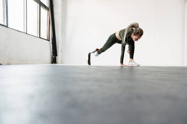 Young sportswoman with prosthesis doing exercise while working out indoors - 写真・画像