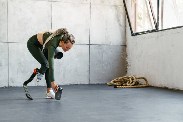Young sportswoman with prosthesis holding water bottle and fitness mat indoors - Foto, Imagen