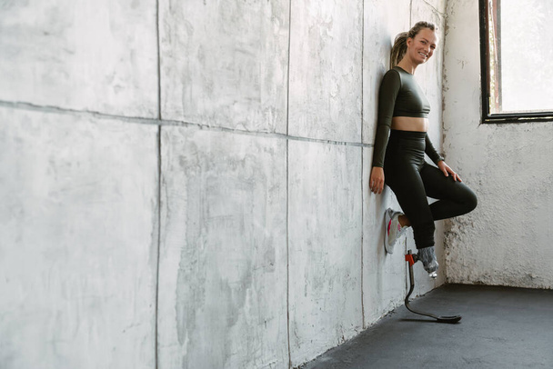 Молодая спортсменка с протезом улыбается, опираясь на стену в помещении - Фото, изображение