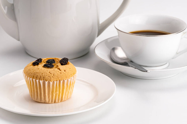 Side view of simply breakfast with raisin sponge cupcake in white ceramic dish and a cup of hot black coffee with metal spoon and a coffee kettle on white background. - Photo, image