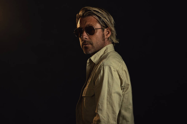 Man with blond hair and a stubble beard and dark aviator sunglasses wears a safari shirt. Looking over his shoulder. - Fotoğraf, Görsel