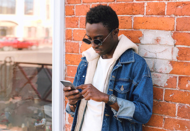 Portrait of modern young african man with smartphone on city street over brick wall background - Foto, imagen