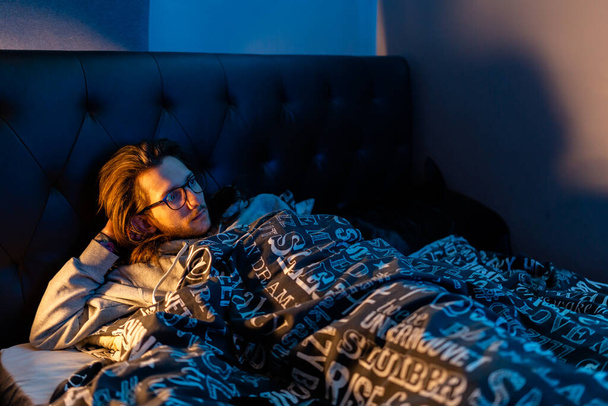 Ένας νεαρός ξαπλωμένος στο κρεβάτι βλέποντας τηλεόραση πριν πάει για ύπνο. Τρόπος ζωής, μετά τη σκοτεινή έννοια - Φωτογραφία, εικόνα