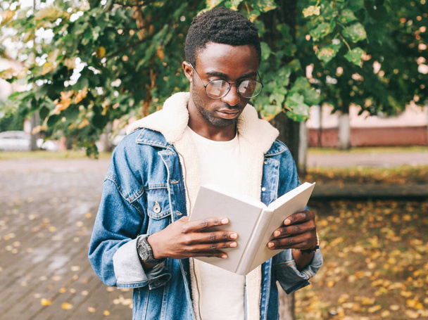 Portrait close up of young african man student reading a book wearing an eyeglasses in autumn city park - Photo, Image