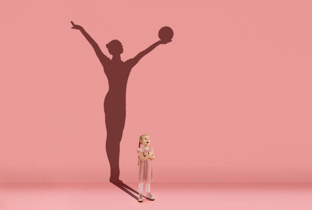 Childhood and dream about big and famous future. Conceptual image with girl and shadow of fit female rhythm gymnast on coral pink background - Фото, изображение