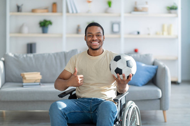 Веселый черный инвалид в инвалидной коляске держит футбольный мяч и показывает большой палец вверх жестом дома - Фото, изображение