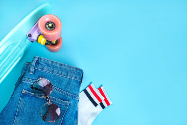 Sport inspired flatlay with turquoise penny skateboard, white striped socks, denim shorts and round grasient sunglasses isolated on blue background. - Photo, Image