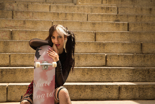 Retrato de una hermosa joven con estilo heterocromo y punk sentada en unas escaleras apoyada en un monopatín que tiene entre las piernas mirando a la cámara. - Foto, Imagen