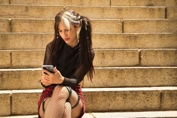 Νέα και όμορφη κοπέλα με κοτσίδες και punk στυλ κάθεται σε κάποια σκάλα συμβουλεύοντας τα κοινωνικά δίκτυα στο κινητό της τηλέφωνο. - Φωτογραφία, εικόνα