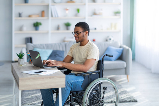 Μαύρος ανάπηρος σε αναπηρική καρέκλα που εργάζεται στο διαδίκτυο από το σπίτι, χρησιμοποιεί φορητό υπολογιστή, ψάχνει για ευκαιρίες εργασίας στο διαδίκτυο - Φωτογραφία, εικόνα