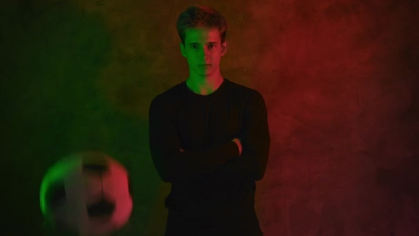 Un apuesto joven futbolista en un jersey negro está de pie con los brazos cruzados sobre su pecho en la luz de neón y una pelota salta delante de él. 4K - Imágenes, Vídeo
