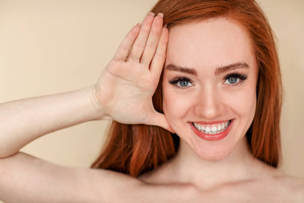 Young ginger female with clean skin touching face and looking at camera with smile against beige background - Foto, Imagen