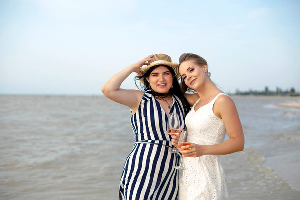 Glad adult female friends in summer outfit smiling while enjoying wine on sandy beach near sea - Фото, зображення