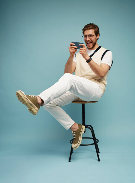 Portret podekscytowanego młodzieńca siedzącego na stołku i grającego w gry na telefonie komórkowym odizolowanego na niebieskim tle. - Zdjęcie, obraz